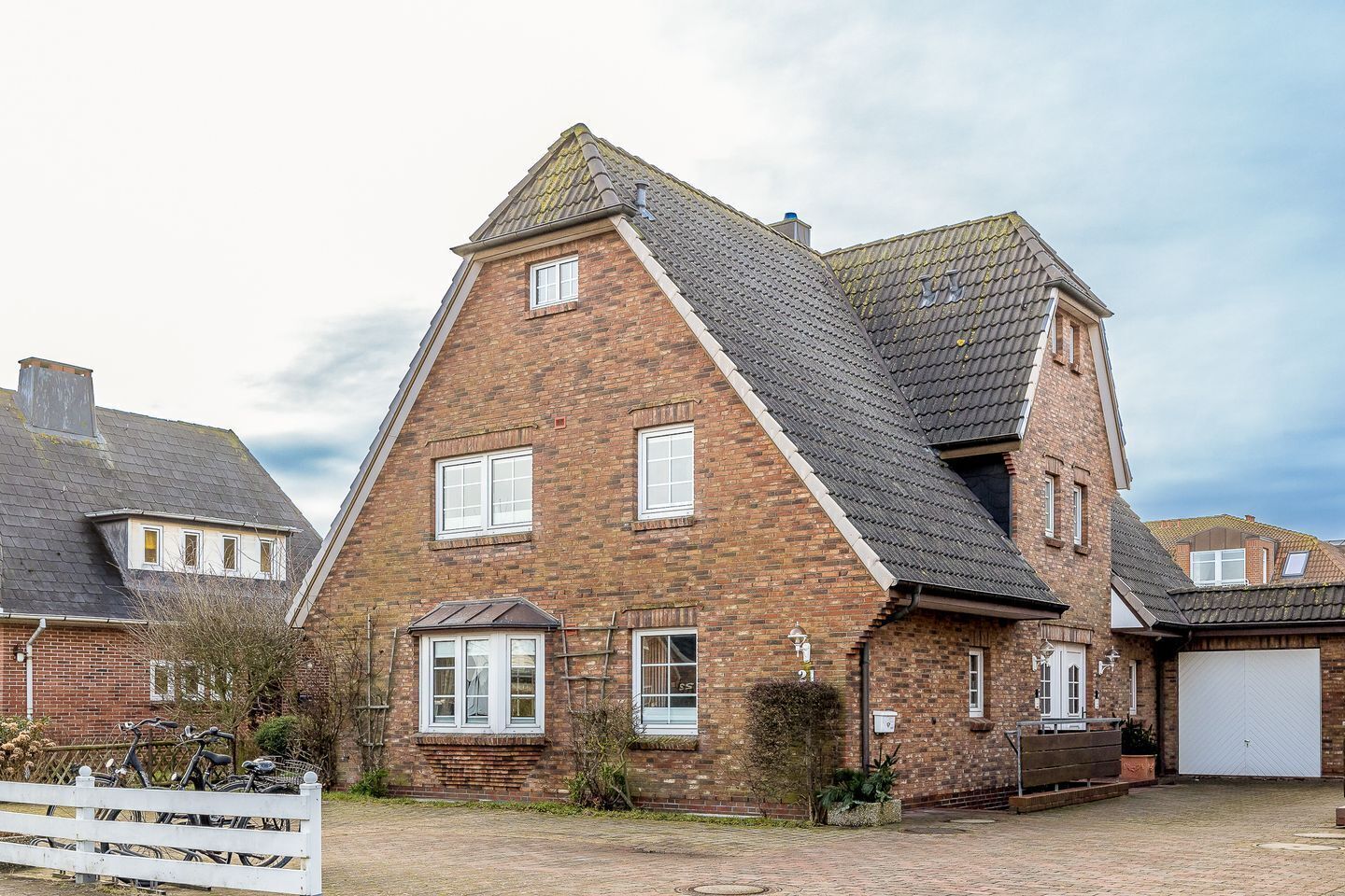 A two-story brick house with a steeply pitched roof, a garage, and a front garden with a white fence and bicycles.