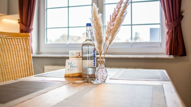 A table with a bottle of MAGNUS, a brochure for Syll Homes by Maja, and pampas grass in a glass vase.