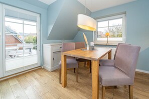 A dining area with a wooden table and chairs, a white pendant lamp, and a view of the outside through large windows. - Appartement Flut No. 7 - Appartement Flut 7 (Sylt)
