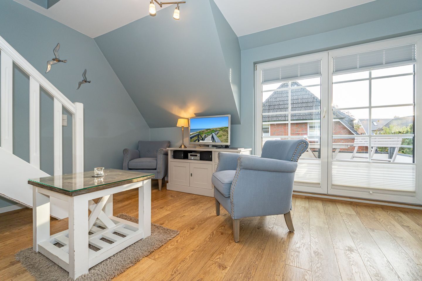 Quarto moderno no estilo loft, com teto inclinado, uma janela grande com persianas, uma TV sobre um móvel branco e uma mesa de vidro.