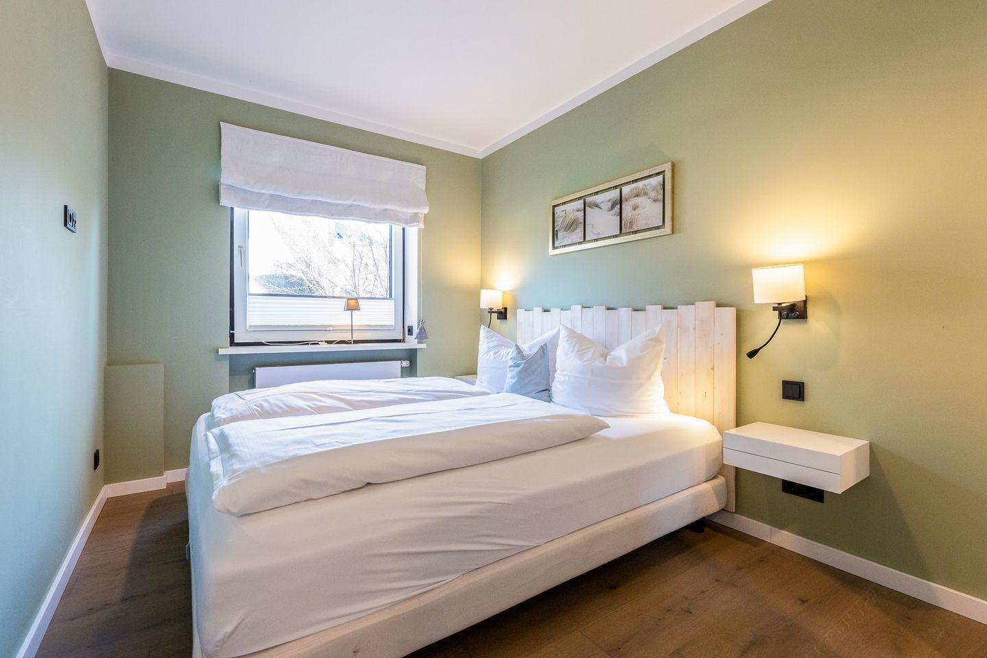 A bedroom with a large bed, a window with a white blind, a bedside table with a lamp, and a wall-mounted light fixture.
