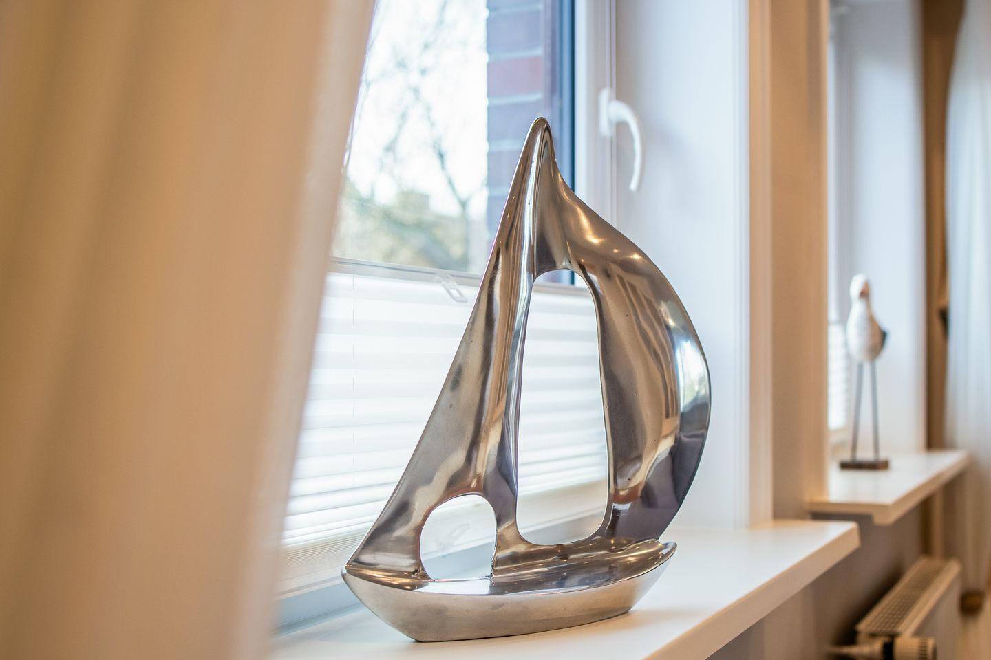 Ein metallisches Segelbootmodell steht auf einem Regal neben einem Fenster mit Jalousien.