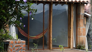 Una terraza de madera con un muro de piedra, una hamaca y una puerta de cristal que da a una habitación con techo de tejas.