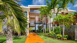 A modern house with a wooden deck, surrounded by tropical plants and palm trees. - Casa Beira-mar com 8 Suítes no Prestigiado Condomínio Iberostar - Praia do Forte (Açu da Torre)