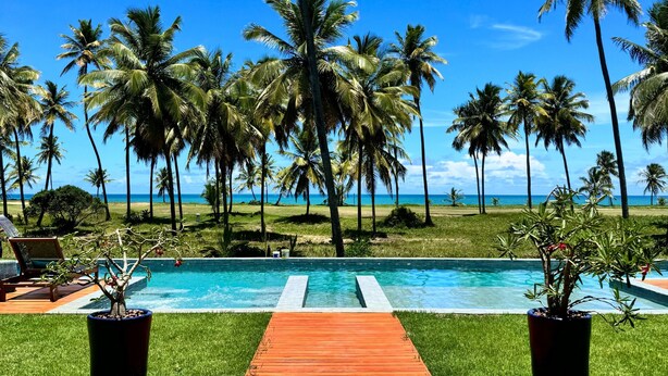 A pool with a wooden deck leading to it, surrounded by palm trees and a view of the ocean. - Casa Beira-mar com 8 Suítes no Prestigiado Condomínio Iberostar - Praia do Forte (Açu da Torre)