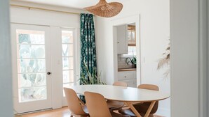 Een eetruimte met een ronde tafel en zes stoelen, een geweven hanglamp en uitzicht op de keuken door een open deur.