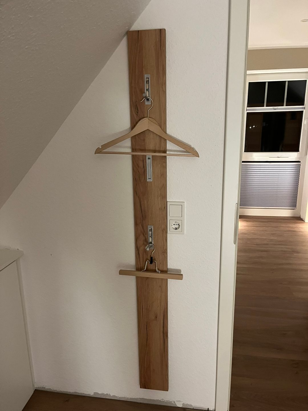 A wooden wall-mounted rack with two hangers and a shelf, positioned in a room with a sloped ceiling and a visible window.