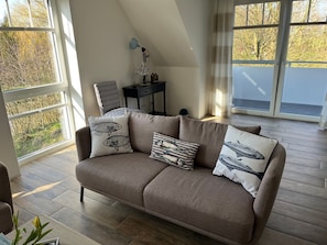 Una sala de estar acogedora con un sofá marrón, cojines estampados, una silla negra y una mesita. Amplias ventanas ofrecen vistas a árboles y una masa de agua.