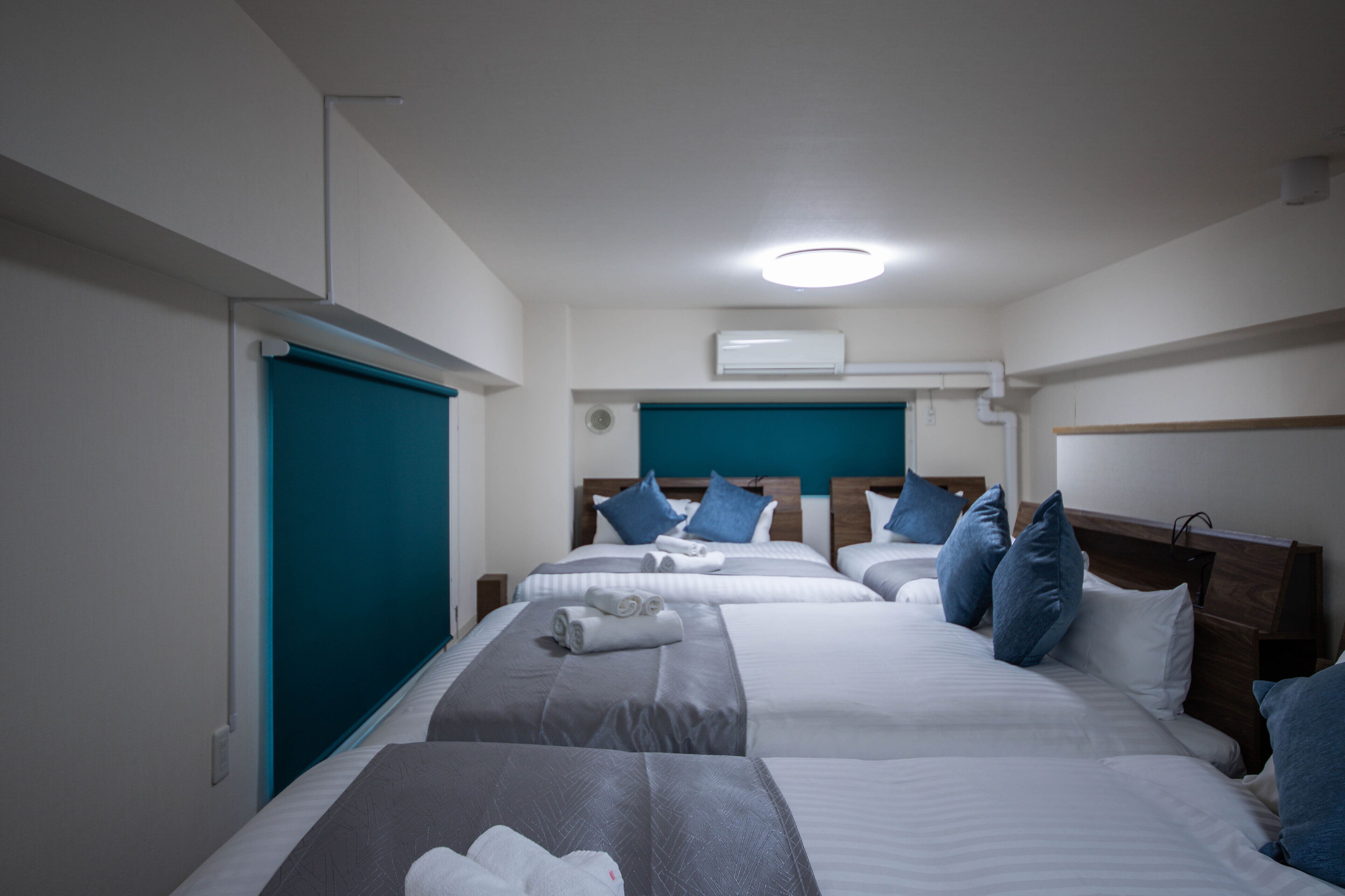Ein Hotelzimmer mit zwei Betten, jeweils mit einem Holz-Kopfteil, weißer Bettwäsche und blauen Kissen. Darüber befinden sich eine Deckenleuchte und eine Klimaanlage.
