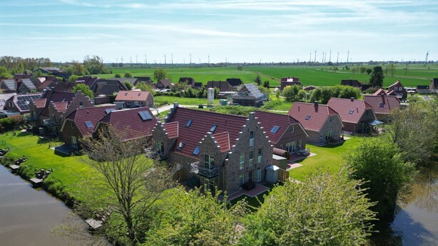Et boligområde med huse, røde tegltage og stenvægge, omgivet af grønne marker og en flod.