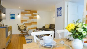 A cozy living room with a wooden floor, a dining table set with plates, glasses, and a bowl, and a chair with a striped cushion.