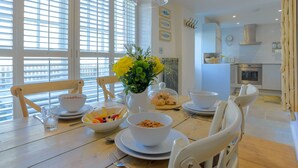 A dining area with a wooden table set for breakfast, including bowls of fruit and cereal, and a vase with yellow flowers.