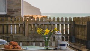 A wooden table with pastries, a coffee pot, and a cup, set against a wooden fence with a view of the sea and cliffs.