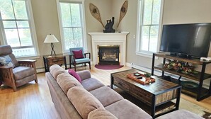 Un salon avec un long canapé beige, une table basse en bois, un téléviseur posé sur un meuble en bois, une cheminée et de grandes fenêtres.