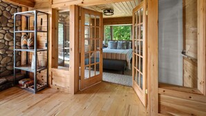 A wooden cabin interior with a stone wall, a metal shelving unit, and a bed with a view of greenery outside. - Mountain Majesty: Timber Lodge with Stunning Views (Morristown)