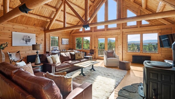 A spacious log cabin interior with a high ceiling, wooden beams, a fireplace, a sofa set, and large windows offering views of the outdoors. - Mountain Majesty: Timber Lodge with Stunning Views (Morristown)