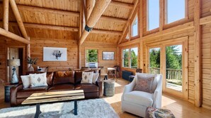 A log cabin interior with a wooden ceiling, a brown leather sofa, a white armchair, and a dining area with a table and chairs. - Mountain Majesty: Timber Lodge with Stunning Views (Morristown)