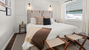 Un dormitorio con una cama grande, dos mesitas de noche, una ventana con cortinas e iluminación en la pared.