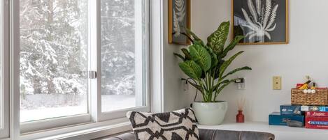 Une pièce avec une grande fenêtre, une plante en pot, un banc rembourré et une étagère fixée au mur, ornée de boîtes et d’une petite bouteille rouge.