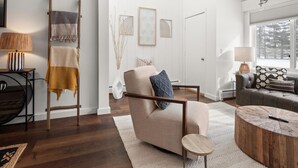 Una sala de estar moderna con un sillón color beige, una mesa de centro redonda y un sofá. Se ve una escalera apoyada en la pared, una lámpara sobre una mesita auxiliar y una ventana con vistas a árboles.