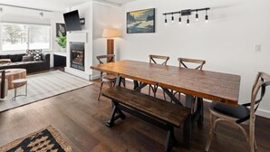 Un comedor con una larga mesa de madera, bancos y sillas. Hay una chimenea, un sofá y una lámpara al fondo.