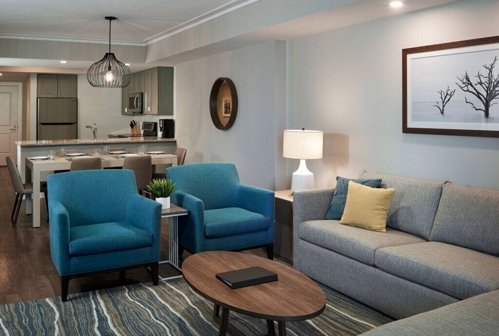 Una sala moderna con un sofá gris, dos sillones azules, una mesa de centro, una zona de comedor con una cocina al fondo y un cuadro enmarcado en la pared.