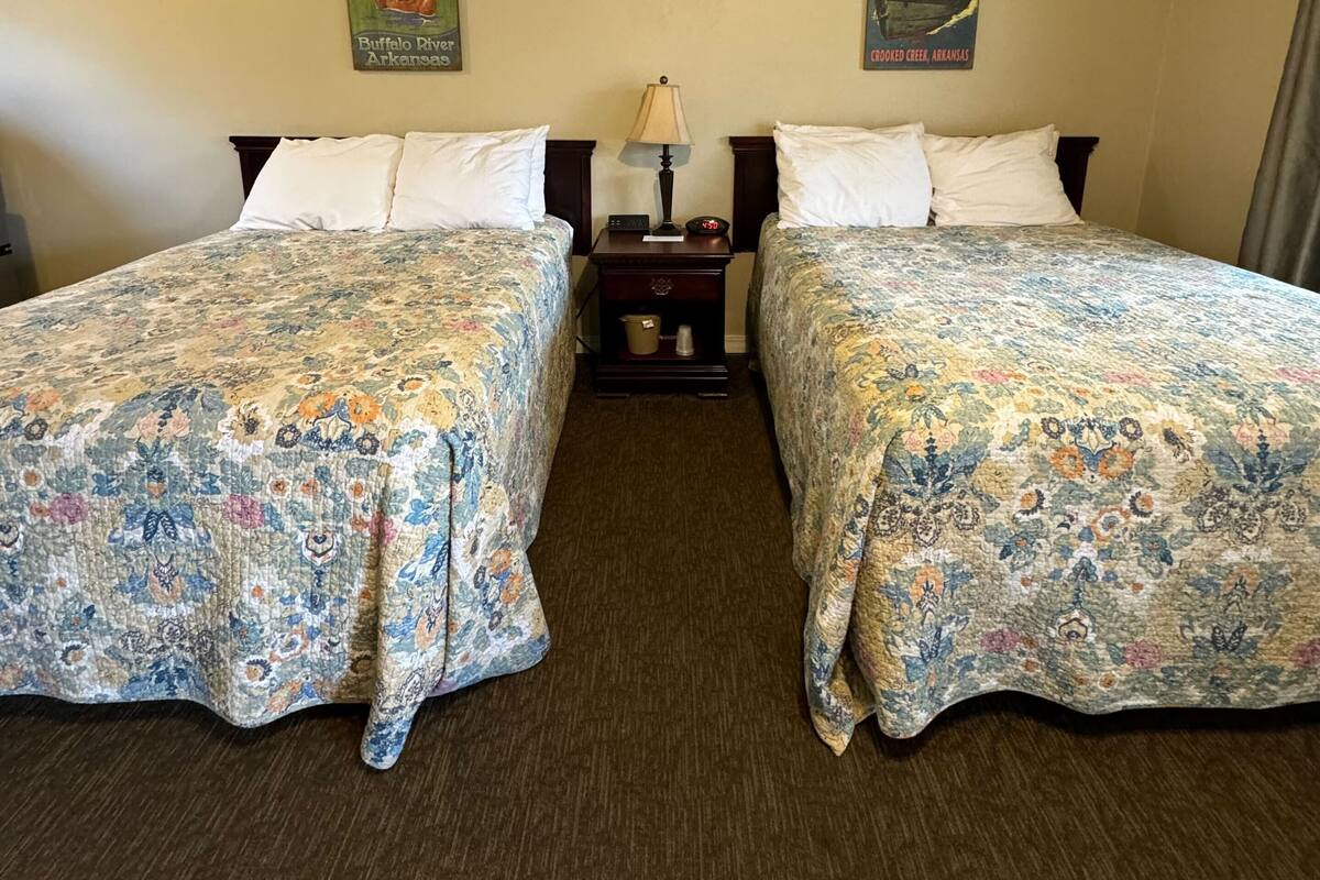 Habitación de hotel con dos camas, colchas estampadas con motivos florales, una mesita de noche con lámpara y dos carteles en la pared.
