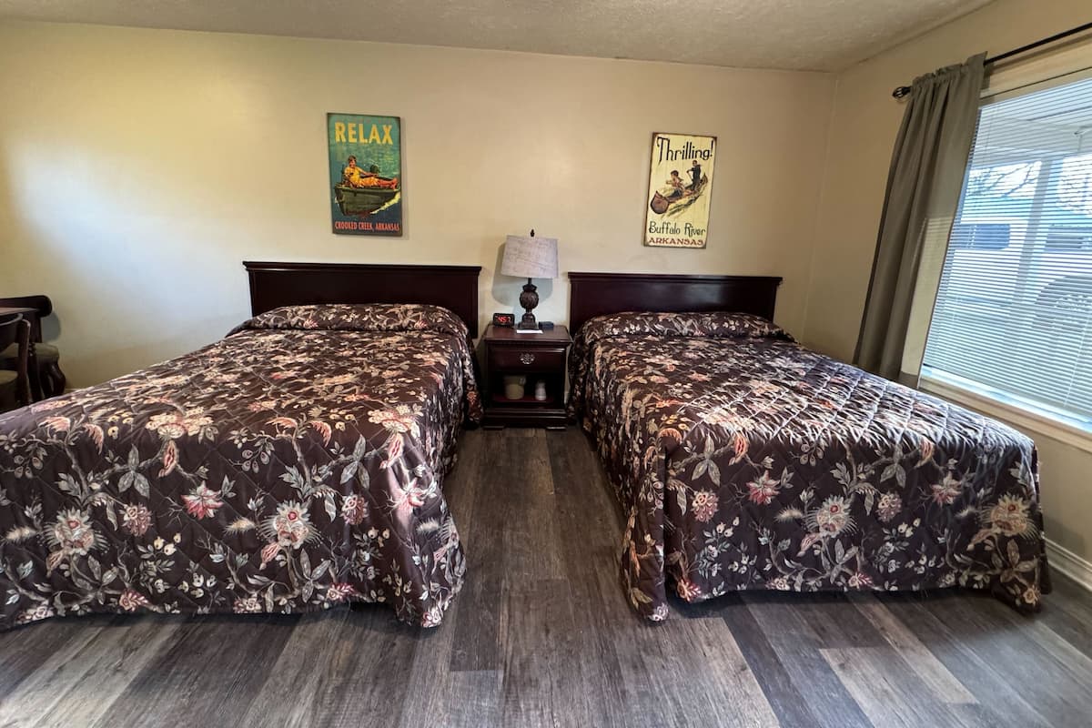 Habitación de hotel con dos camas, cabeceros de madera, cubrecamas florales, una mesita de noche con lámpara y dos carteles en la pared.