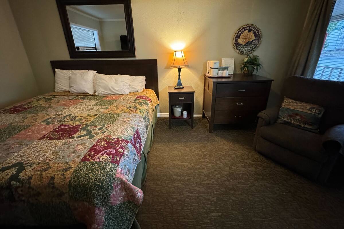 Habitación de hotel con una cama grande, una mesita de noche con lámpara, cómoda, silla y un reloj de pared.