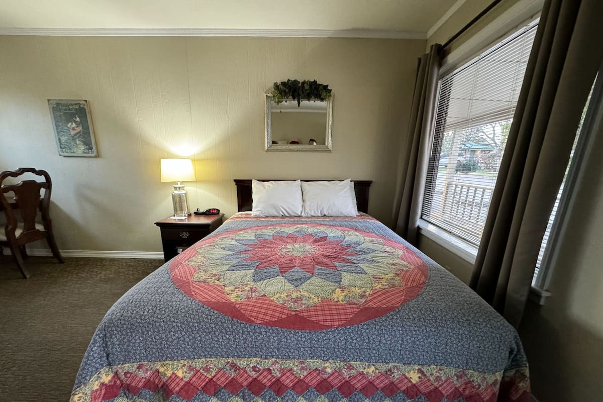 Una cama bien hecha con un edredón colorido, una lámpara de noche, una silla, una mesita con un reloj y una ventana con cortinas.