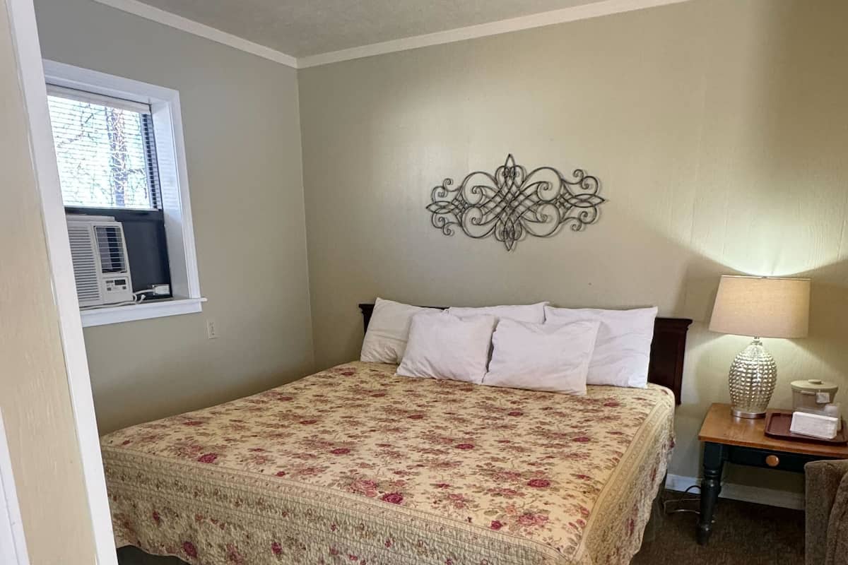 Un dormitorio con cama, ventana con aire acondicionado, un adorno en la pared, lámpara de noche y mesita de noche con lámpara y bandeja.