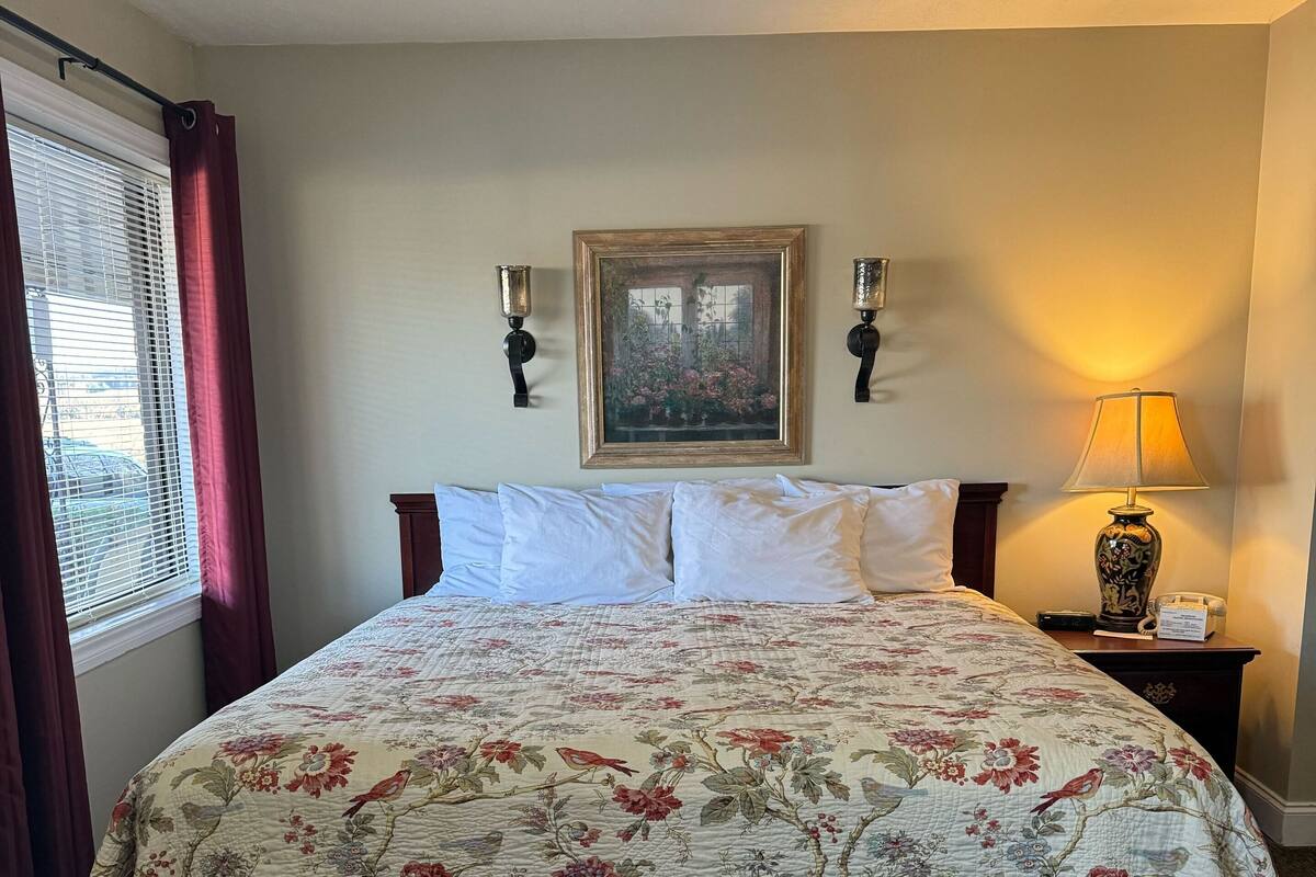 Una cama bien hecha con una colcha estampada y falda a rayas, un cuadro en la pared, dos apliques, una lámpara de noche y una ventana con cortinas.