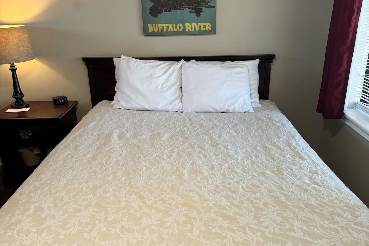 Una cama bien tendida con una colcha blanca y dos almohadas, un reloj de pared y un cartel enmarcado sobre la cama.
