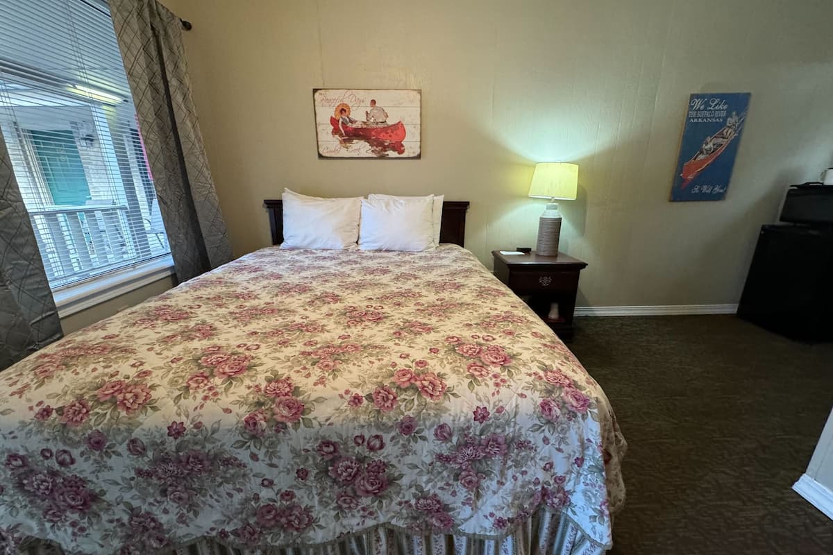 Habitación de hotel con una colcha estampada con motivos florales, una mesita de noche con una lámpara, un cuadro enmarcado en la pared y una ventana con cortinas.