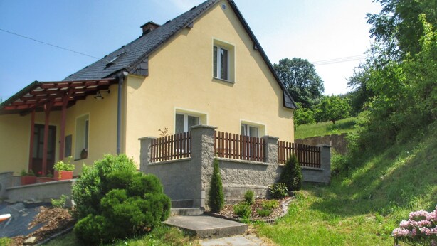 Une maison à deux  étages avec une façade en pierre, une terrasse en bois et un chemin en pierre menant à l’entrée.