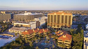 Un complejo hotelero con varias edificaciones, incluyendo un hotel grande con techo rojo y una edificación más pequeña con fachada blanca. Hay una zona de piscina con palmeras y vistas de la ciudad circundante.