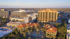 Un complexe hôtelier comprenant plusieurs bâtiments, dont un grand hôtel avec un toit rouge et un bâtiment plus petit à la façade blanche. On y trouve un espace piscine agrémenté de palmiers et offrant une vue sur le paysage urbain environnant.