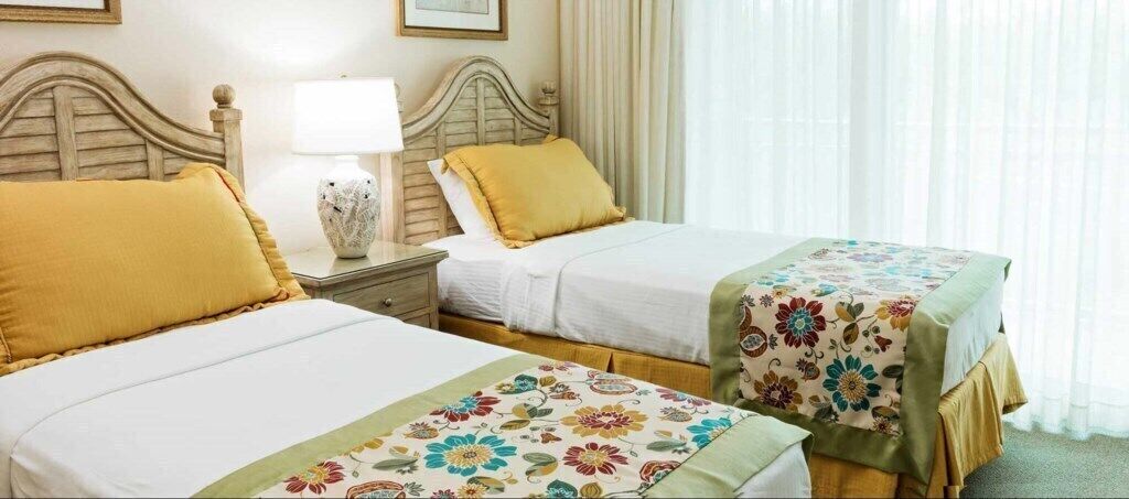Een hotelkamer met twee bedden, elk met een ander bloemenbedekking, een nachtkastje met lamp en een raam met dunne gordijnen.