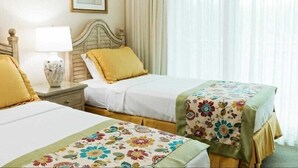 Ein Hotelzimmer mit zwei Betten, jedes mit einer unterschiedlichen Blumendekoration, einem Nachttisch mit Lampe und einem Fenster mit transparenten Vorhängen.