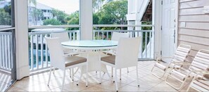 A covered outdoor area with a glass table and white chairs, a white railing, and a view of greenery and buildings. - Sanibel Island | Relaxing Coastal Retreat | Pool + Beach | 2BR (Sanibel Island)