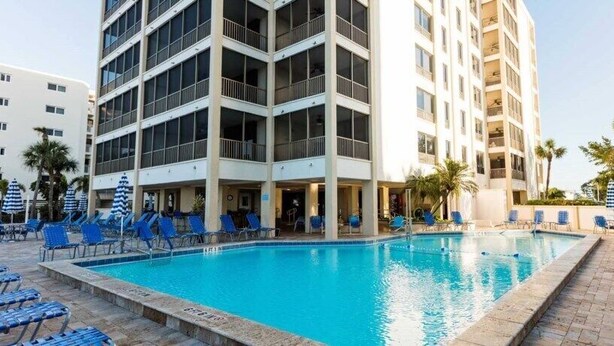 Un espace piscine d’hôtel doté de plusieurs niveaux de balcons, de chaises longues bleues et d’une piscine à l’eau d’un bleu limpide.