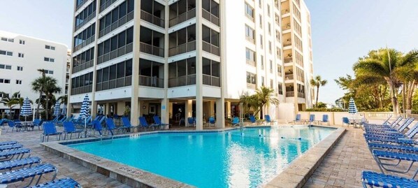 Zona de piscina de hotel con varios niveles de balcones, sillas reclinables azules y una piscina de color azul cristalino.