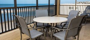 A round table with four chairs, a balcony with a metal railing, and a view of the beach and city skyline.