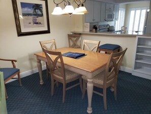A dining area with a wooden table and chairs, a framed picture on the wall, and a kitchen area with cabinets and appliances.