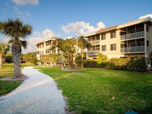 A pathway leading to a multi-story building with balconies, surrounded by well-maintained grass and palm trees.