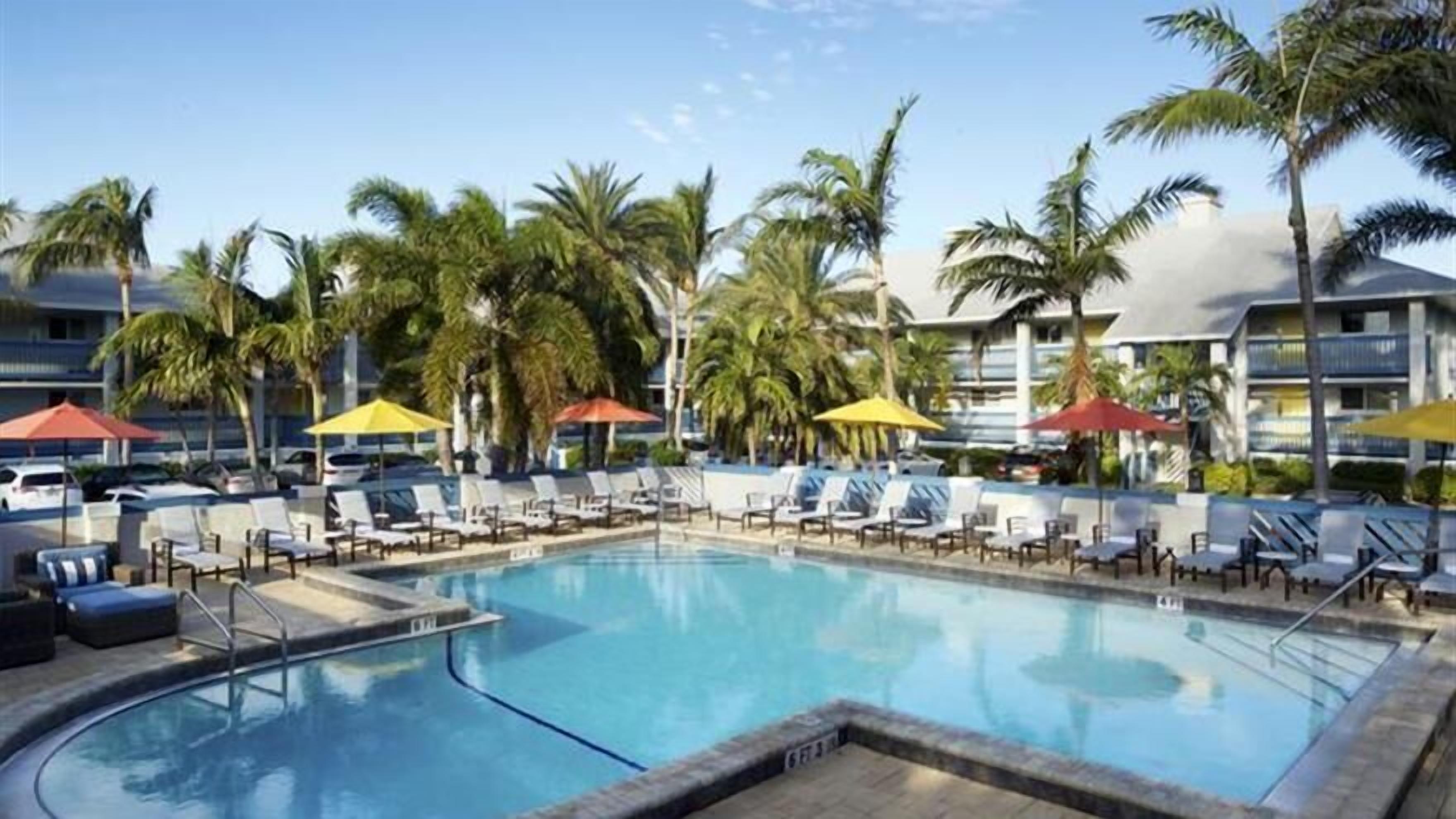 Une piscine entourée de palmiers et de bâtiments d’hôtel, avec des chaises longues et des parasols.