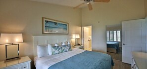 Une chambre à coucher comprenant un lit, des tables de chevet, un ventilateur de plafond, un tableau et une porte donnant sur une autre pièce.