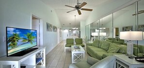 Ein modernes Wohnzimmer mit Fernseher, grünen Möbeln, Deckenventilator und einer Küche im Hintergrund.