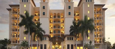 A multi-story hotel with a central entrance, palm trees, and a well-lit facade.