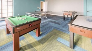Una mesa de billar con superficie de fieltro verde, una mesa de fútbol de mesa y una mesa de ping pong en una sala con piso de alfombra estampada.